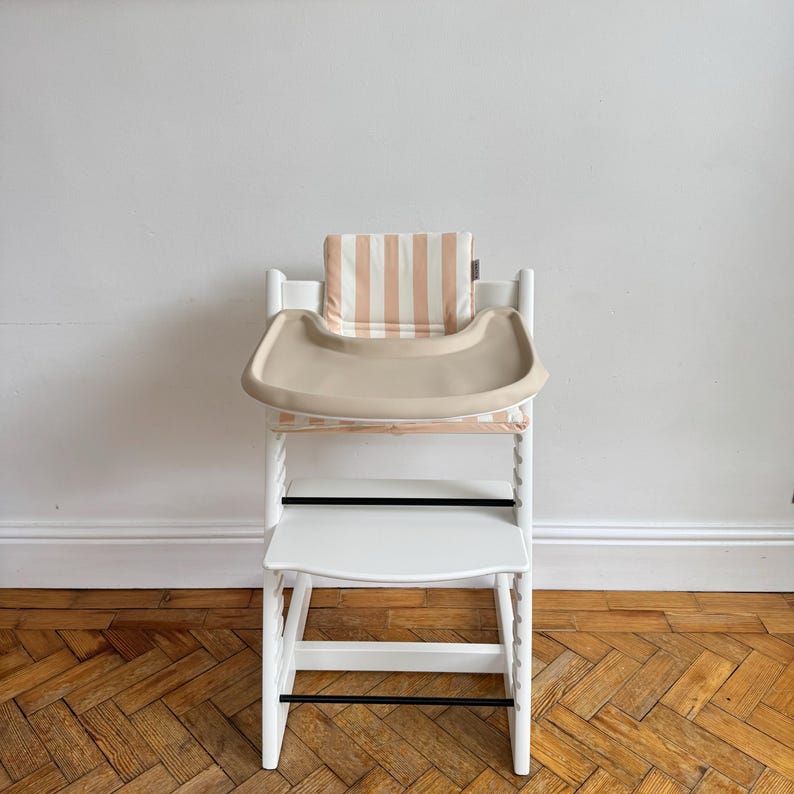 Pu&ograve; includere: Un seggiolone in legno bianco con vassoio in plastica beige e cuscino imbottito a righe verticali color pesca chiaro e bianco. La sedia presenta una struttura con barre di supporto nere orizzontali e un design del sedile regolabile.