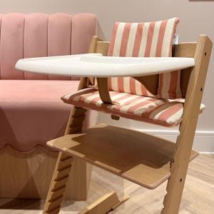 Pu&ograve; includere: Un seggiolone in legno con vassoio in plastica bianca e cuscino di seduta a righe verticali color corallo e bianco. La sedia include un cuscino per lo schienale a righe abbinato con una piccola etichetta bianca con la scritta cove baby.
