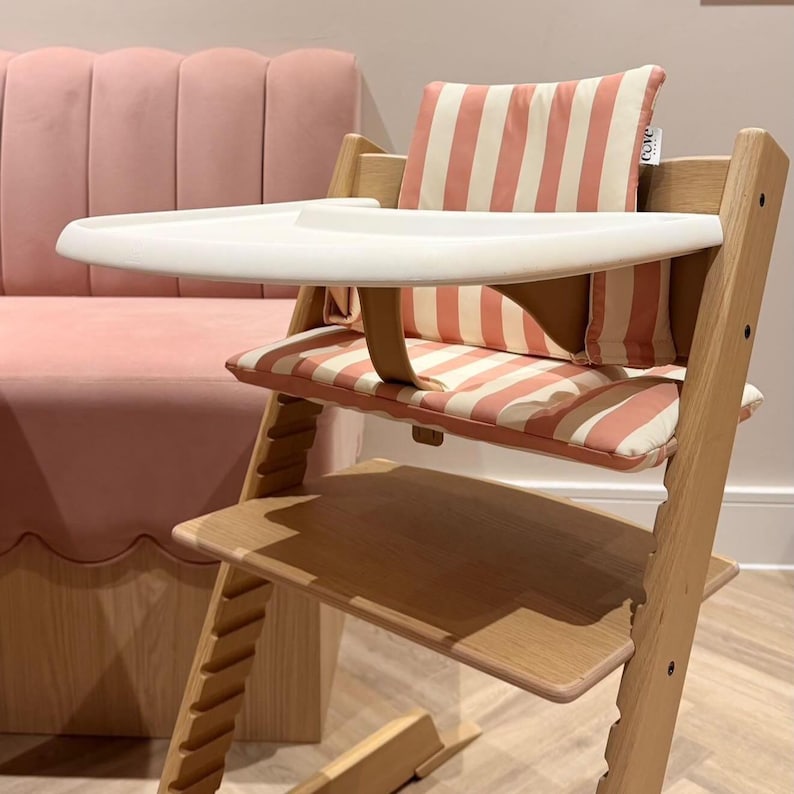 Pu&ograve; includere: Un seggiolone in legno con vassoio in plastica bianca e cuscino di seduta a righe verticali color corallo e bianco. La sedia include un cuscino per lo schienale a righe abbinato con una piccola etichetta bianca con la scritta cove baby.