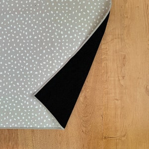 Könnte beinhalten: Ein grauer Teppich mit einem weißen Tupfenmuster. Die Unterseite des Teppichs ist schwarz und strukturiert. Der Teppich ist teilweise aufgerollt und zeigt die schwarze Unterseite und den Holzboden.