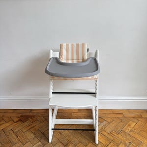 Pu&ograve; includere: Un seggiolone in legno bianco dotato di vassoio rimovibile in plastica grigia e cuscino imbottito con motivo a righe verticali beige e bianche. La sedia include un poggiapiedi e una base di seduta con barre di supporto orizzontali nere.