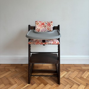 May include: A dark brown wooden high chair featuring a grey plastic tray and a padded seat cushion with a colourful abstract floral pattern in shades of red, orange, and purple. The chair includes a matching backrest cushion and a footrest.