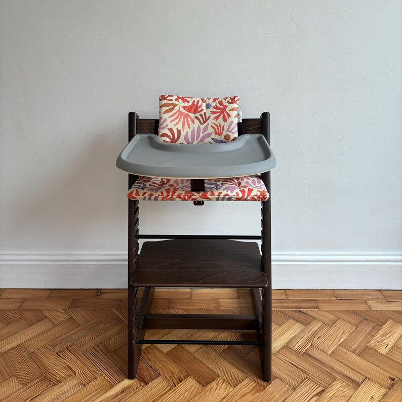 May include: A dark brown wooden high chair featuring a grey plastic tray and a padded seat cushion with a colourful abstract floral pattern in shades of red, orange, and purple. The chair includes a matching backrest cushion and a footrest.