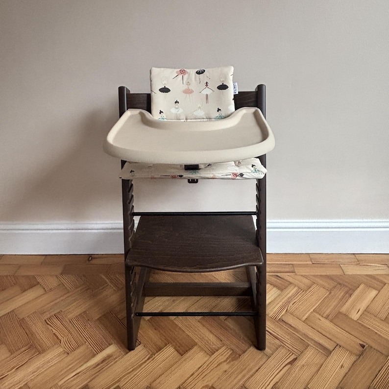 May include: A dark brown wooden high chair featuring a beige plastic tray and a cushioned seat and backrest pad. The fabric cushions are decorated with a repeating pattern of small, stylized ballerinas in shades of pink, black, and teal.