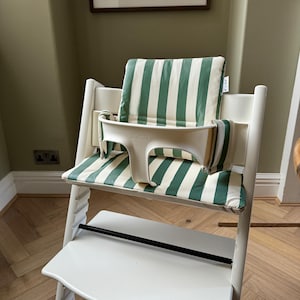 Puede incluir: Silla alta blanca con cojín y respaldo a rayas verdes y blancas. La silla tiene una bandeja blanca y un reposapiés negro. La marca "Stokke" es visible en la bandeja.