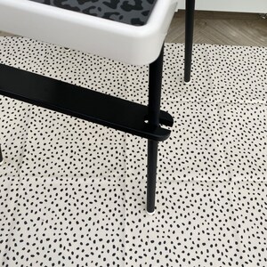 A black wooden footrest that is fitted on a toddlers  high chair legs. Accompanied with a black panther spotted place mat in the tray, black leg wraps and on top of a black spotted pink splash mat.