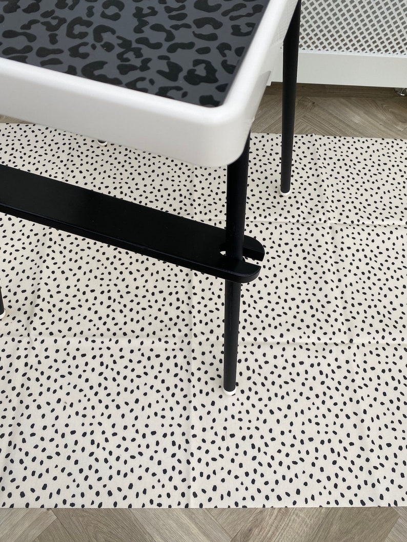 A black wooden footrest that is fitted on a toddlers  high chair legs. Accompanied with a black panther spotted place mat in the tray, black leg wraps and on top of a black spotted pink splash mat.
