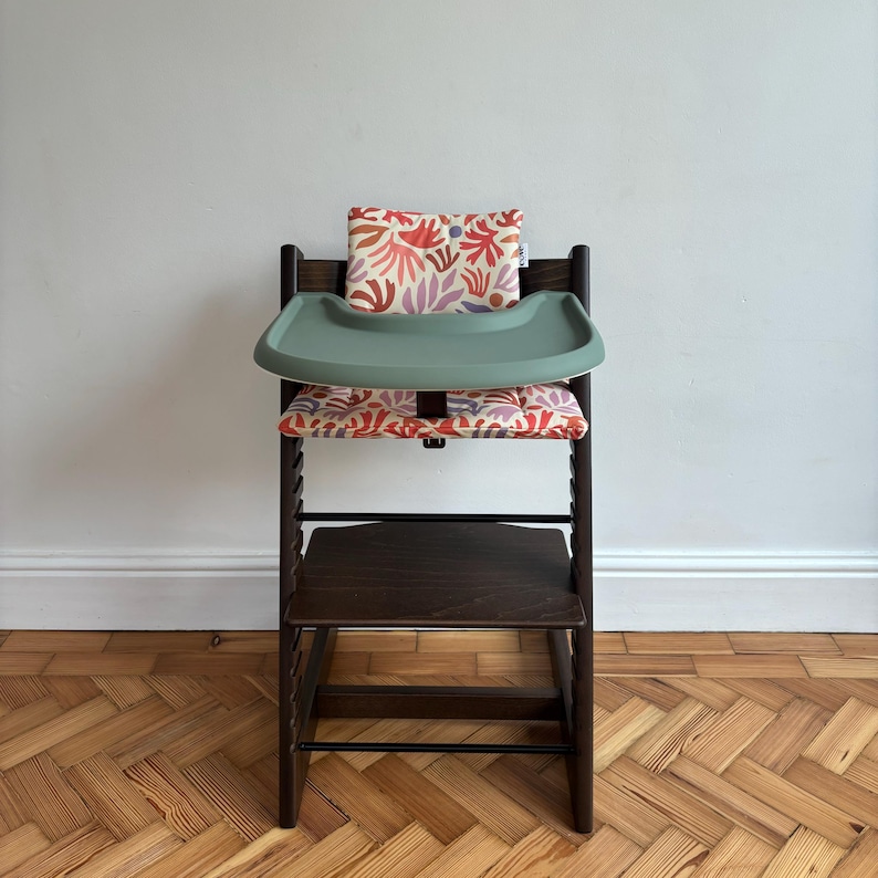 May include: A dark brown wooden high chair featuring a sage green plastic tray and a padded seat cushion with a colourful abstract botanical print in shades of red, orange, and purple. The chair includes a matching backrest cushion.