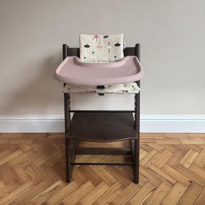 May include: A dark brown wooden high chair featuring a removable dusty pink plastic tray. The chair includes a cream-colored fabric cushion set with a repeating pattern of small illustrated ballerinas in various poses and colors.