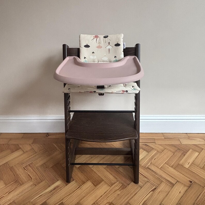 May include: A dark brown wooden high chair featuring a removable dusty pink plastic tray. The chair includes a cream-colored fabric cushion set with a repeating pattern of small illustrated ballerinas in various poses and colors.