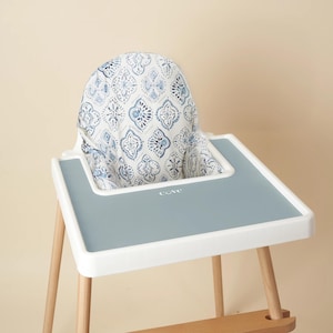 Puede incluir: Una silla alta blanca con una funda de asiento estampada en azul y blanco. La silla tiene una bandeja gris con la palabra "COVE" impresa en ella. La silla tiene patas de madera.