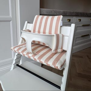 Pu&ograve; includere: Un seggiolone in legno bianco dotato di cuscino imbottito per seduta e schienale con un motivo a righe verticali rosa polvere e bianco. La sedia include un attacco di sicurezza in plastica bianca e una barra poggiapiedi orizzontale nera.