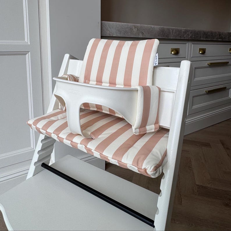 Pu&ograve; includere: Un seggiolone in legno bianco dotato di cuscino imbottito per seduta e schienale con un motivo a righe verticali rosa polvere e bianco. La sedia include un attacco di sicurezza in plastica bianca e una barra poggiapiedi orizzontale nera.
