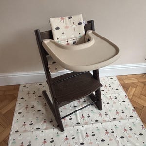 May include: A dark brown wooden high chair featuring a beige plastic tray and a cushioned backrest with a repeating pattern of stylized dancers in pink, black, and teal. The chair includes a matching floor mat with the same dancer print.