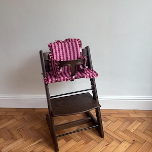 Pode incluir: Uma cadeira alta de madeira marrom escuro com uma almofada e capa de assento listradas em rosa e roxo. A almofada tem uma borda com babados. A cadeira alta está em um piso de madeira.