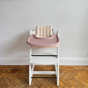 Pu&ograve; includere: Un seggiolone in legno bianco dotato di un vassoio in plastica color rosa polvere e un cuscino imbottito con un motivo a righe verticali beige e bianche. La sedia include un poggiapiedi bianco e due sottili barre di supporto orizzontali nere.