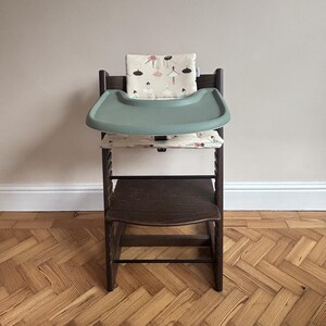 May include: A dark brown wooden high chair featuring a sage green plastic tray and a cream-colored fabric cushion set decorated with a repeating pattern of small ballerinas in various poses, including black, pink, and teal colored outfits.