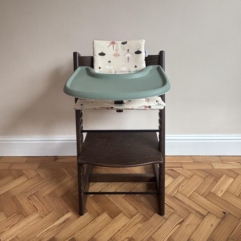 May include: A dark brown wooden high chair featuring a sage green plastic tray and a cream-colored fabric cushion set decorated with a repeating pattern of small ballerinas in various poses, including black, pink, and teal colored outfits.