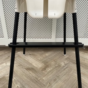 A black wooden footrest that is fitted on a toddlers  high chair legs. Accompanied with black leg wraps on the high chair legs. Placed on a wooden floor.