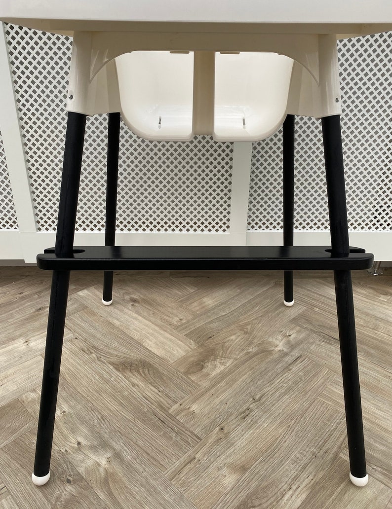 A black wooden footrest that is fitted on a toddlers  high chair legs. Accompanied with black leg wraps on the high chair legs. Placed on a wooden floor.