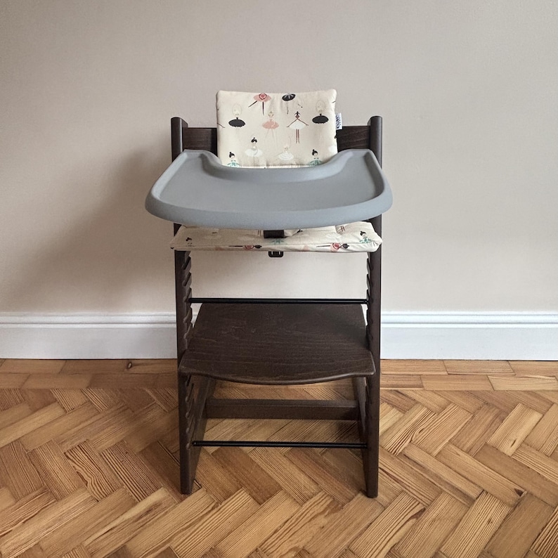 May include: A dark brown wooden high chair featuring a removable gray plastic tray. The chair includes a padded backrest and seat cushion, both decorated with a repeating pattern of small ballerinas in black, pink, and white on a cream background.