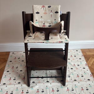 May include: A dark brown wooden high chair featuring a padded seat and backrest cushion with a cream-colored fabric printed with small, colorful ballerina figures. The chair includes a dark brown plastic safety guard and a matching floor mat.