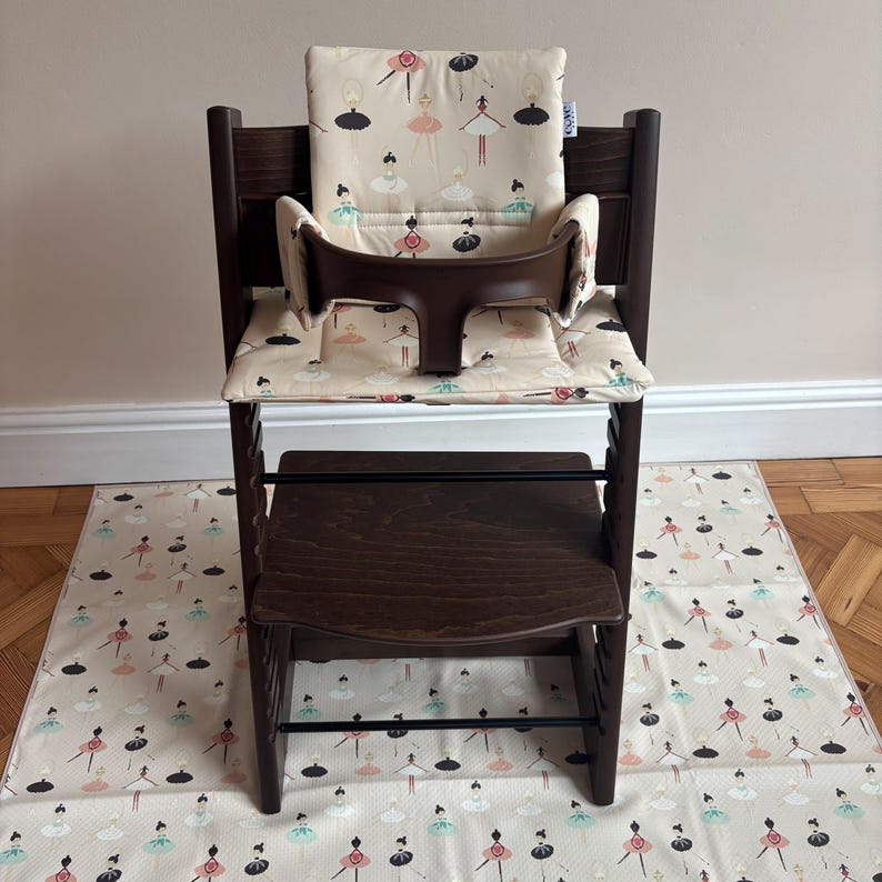 May include: A dark brown wooden high chair featuring a padded seat and backrest cushion with a cream-colored fabric printed with small, colorful ballerina figures. The chair includes a dark brown plastic safety guard and a matching floor mat.