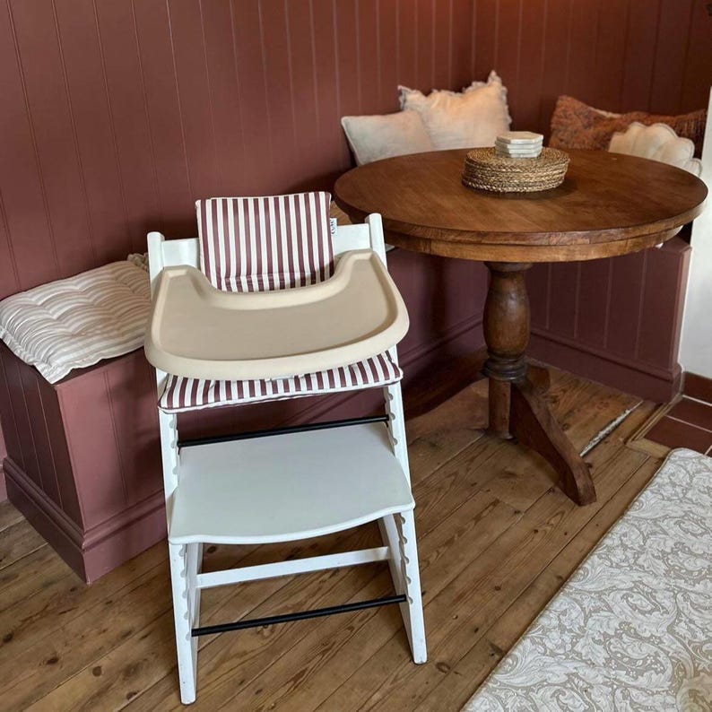 A high chair with a cream full cover silicone place mat on the attached tray. Also includes a brown and white vertical striped cushion in the seat of a chair. Placed next to a wooden table on a wooden floor.