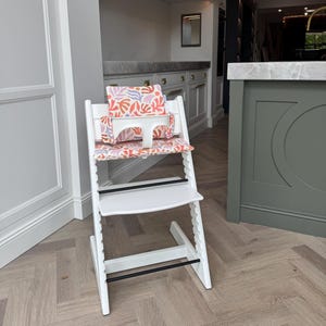 May include: A white wooden high chair featuring a seat and backrest cushion with a colourful abstract pattern of coral, purple, and light pink organic shapes. The chair includes a white plastic safety bar and two black horizontal support bars.