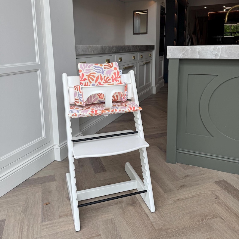May include: A white wooden high chair featuring a seat and backrest cushion with a colourful abstract pattern of coral, purple, and light pink organic shapes. The chair includes a white plastic safety bar and two black horizontal support bars.