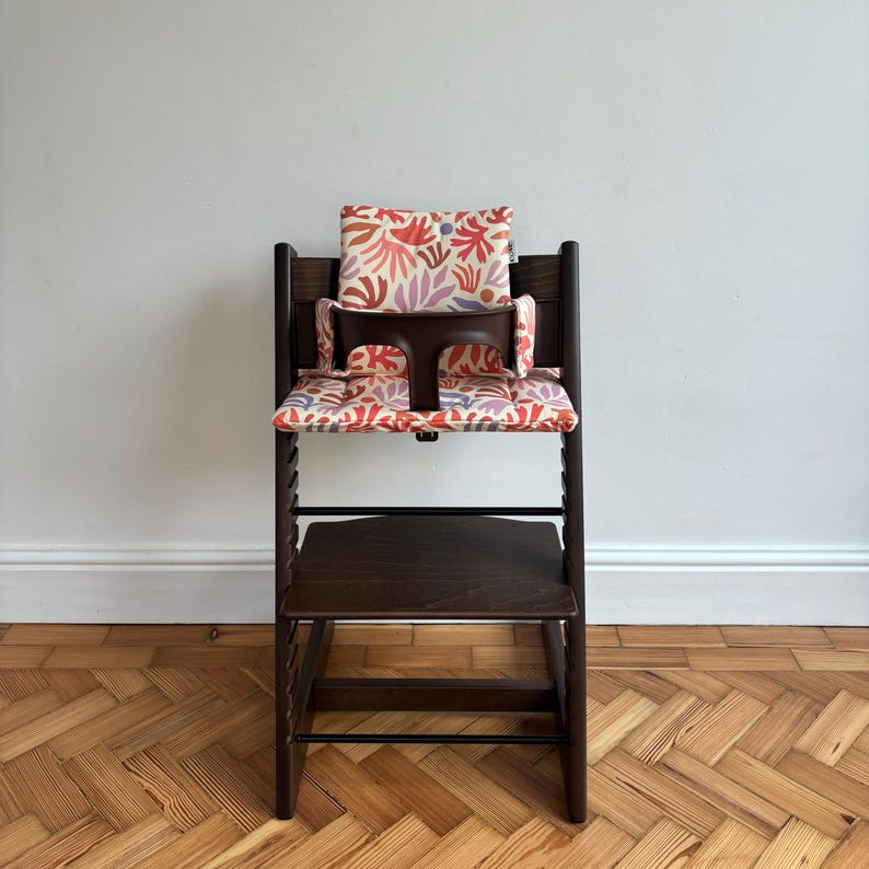 May include: A dark brown wooden high chair featuring a cushioned seat and backrest pad with a colourful abstract botanical pattern in shades of red, pink, and light purple. The chair includes a dark brown plastic safety bar and adjustable footrest.