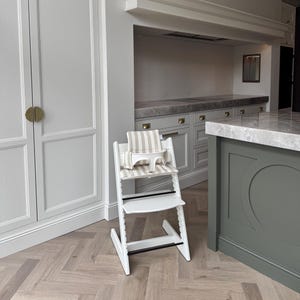 May include: A white wooden high chair with a beige and white striped cushion and safety harness. The high chair has a footrest and a tray. The chair is in a kitchen setting with a light-colored wood floor and white cabinets.
