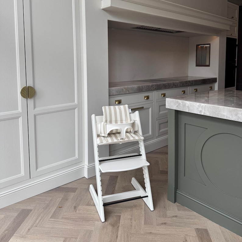May include: A white wooden high chair with a beige and white striped cushion and safety harness. The high chair has a footrest and a tray. The chair is in a kitchen setting with a light-colored wood floor and white cabinets.