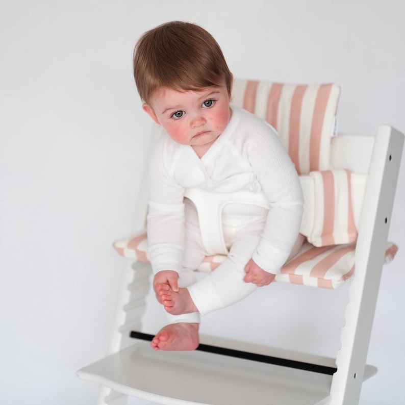 Pu&ograve; includere: Un seggiolone in legno bianco dotato di un cuscino imbottito per la seduta e lo schienale con un motivo a righe verticali rosa chiaro e bianco. La sedia include una cintura di sicurezza in plastica bianca e un poggiapiedi per il bambino.