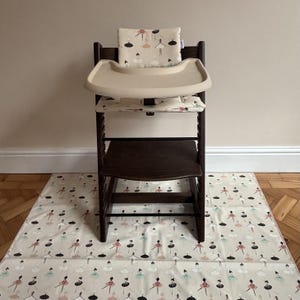 May include: A dark brown wooden high chair featuring a beige tray and a cushioned seat pad with a repeating pattern of ballerinas in pink, black, and teal. A matching floor mat with the same ballerina print is placed underneath the chair.