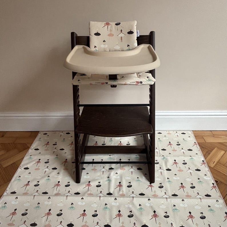 May include: A dark brown wooden high chair featuring a beige tray and a cushioned seat pad with a repeating pattern of ballerinas in pink, black, and teal. A matching floor mat with the same ballerina print is placed underneath the chair.