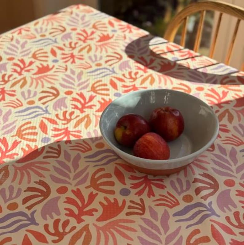 May include: A white ceramic bowl with a terracotta base containing three red apples, resting on a tablecloth featuring a repeating pattern of abstract coral-like shapes and dots in shades of orange, light purple, and pink against a cream background.
