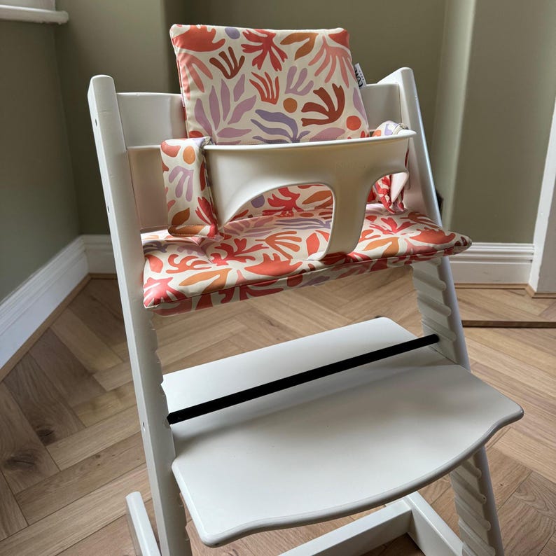 May include: A white wooden high chair featuring a removable cream-coloured plastic safety rail and a cushioned seat pad and backrest. The fabric displays a repeating abstract pattern in shades of orange, light purple, and cream throughout the set.