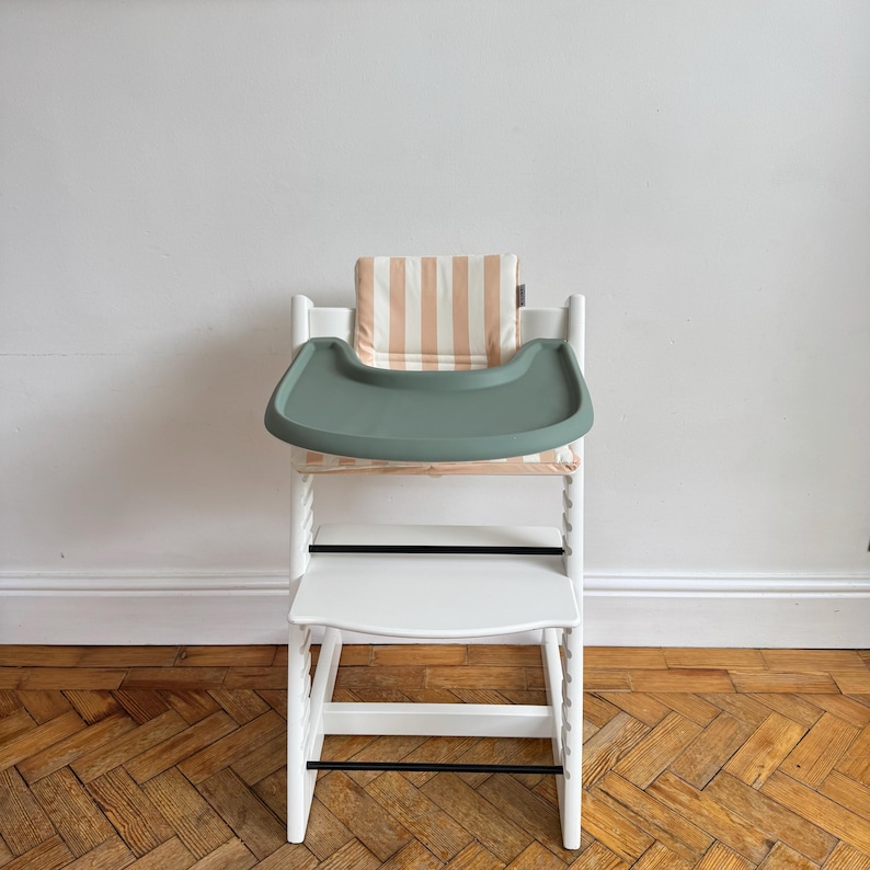 Pu&ograve; includere: Un seggiolone in legno bianco dotato di un vassoio in plastica verde salvia e di un cuscino imbottito con un motivo a righe verticali beige e bianche. La sedia include un poggiapiedi, una seduta e due sottili barre di supporto orizzontali nere.