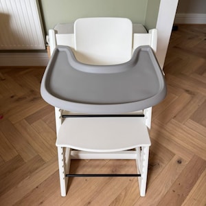 May include: A white high chair with a gray tray and footrest. The chair has a modern design with black support bars and is made of wood and plastic. The chair is designed for a baby or toddler.