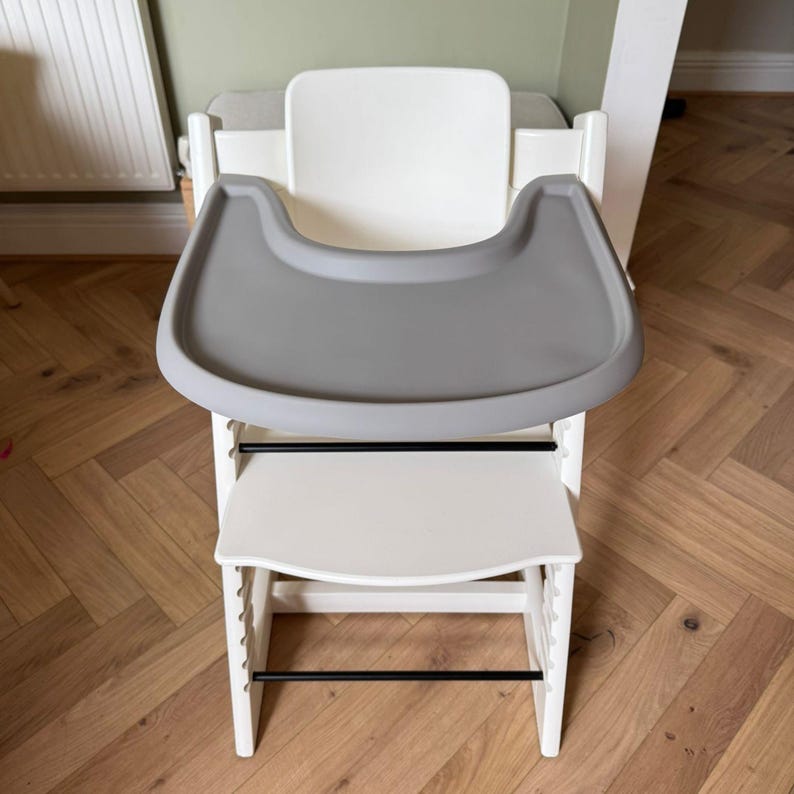 May include: A white high chair with a gray tray and footrest. The chair has a modern design with black support bars and is made of wood and plastic. The chair is designed for a baby or toddler.