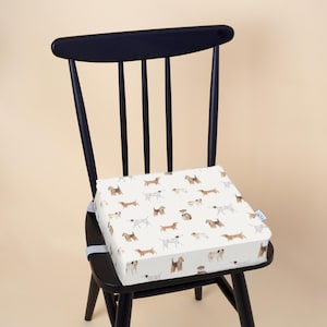 May include: A black wooden chair with a square cushion featuring a repeating pattern of various dog breeds in brown, black, and white. The cushion is secured to the chair with a white strap. The chair is set against a neutral background.