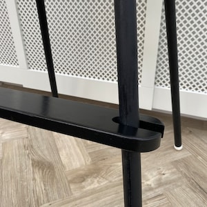 A close up of a black wooden footrest that is fitted on a toddlers  high chair legs. Accompanied with black leg wraps on the high chair legs, placed on a wooden floor.