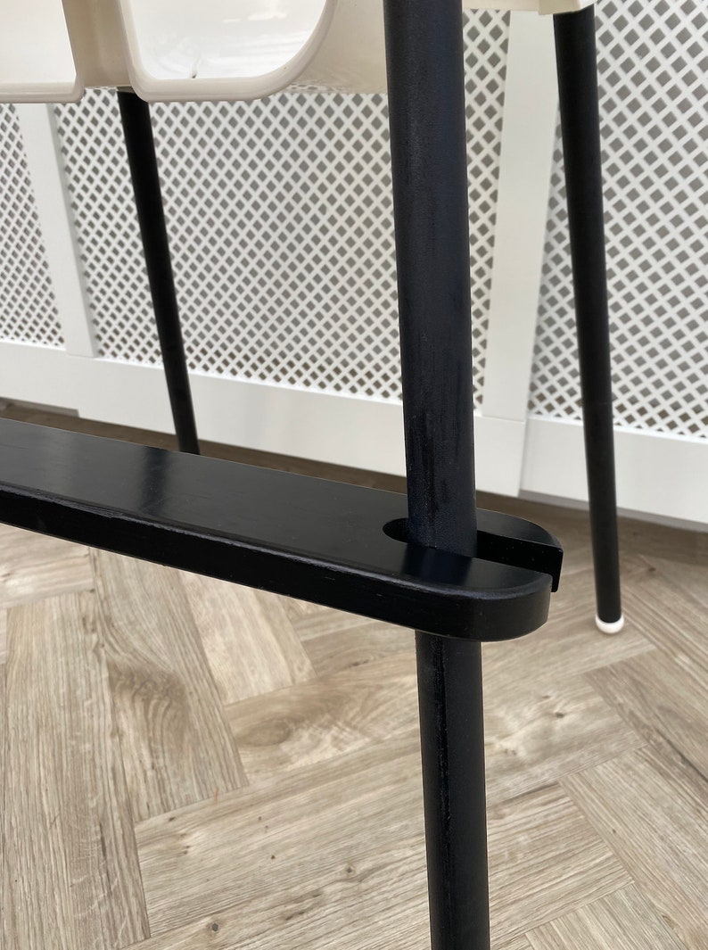 A close up of a black wooden footrest that is fitted on a toddlers  high chair legs. Accompanied with black leg wraps on the high chair legs, placed on a wooden floor.