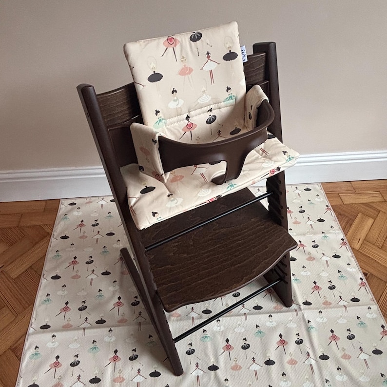May include: A dark brown wooden high chair featuring a beige padded seat cushion and backrest decorated with a repeating pattern of stylized ballerinas in black, pink, and teal. The chair includes a dark brown plastic safety guard and footrest.