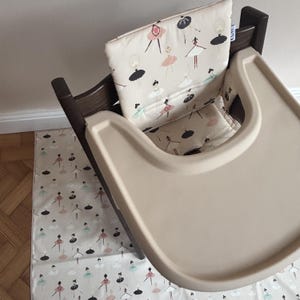 May include: A dark brown wooden high chair featuring a beige padded seat cushion and a matching beige floor mat. Both the cushion and the mat are decorated with a repeating pattern of ballerinas in black, pink, and light blue tutus.