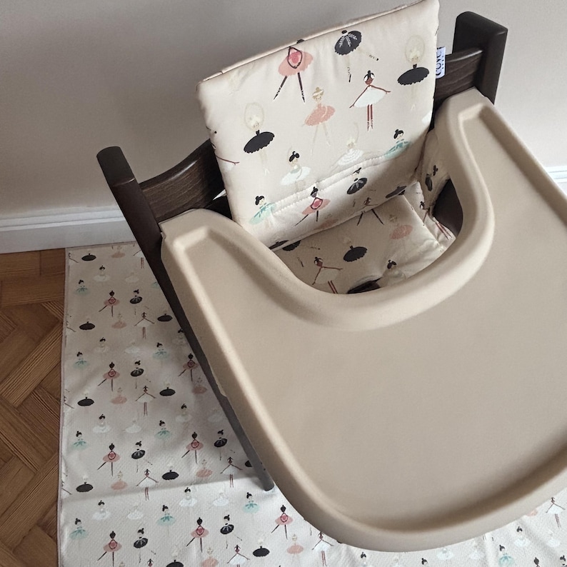 May include: A dark brown wooden high chair featuring a beige padded seat cushion and a matching beige floor mat. Both the cushion and the mat are decorated with a repeating pattern of ballerinas in black, pink, and light blue tutus.
