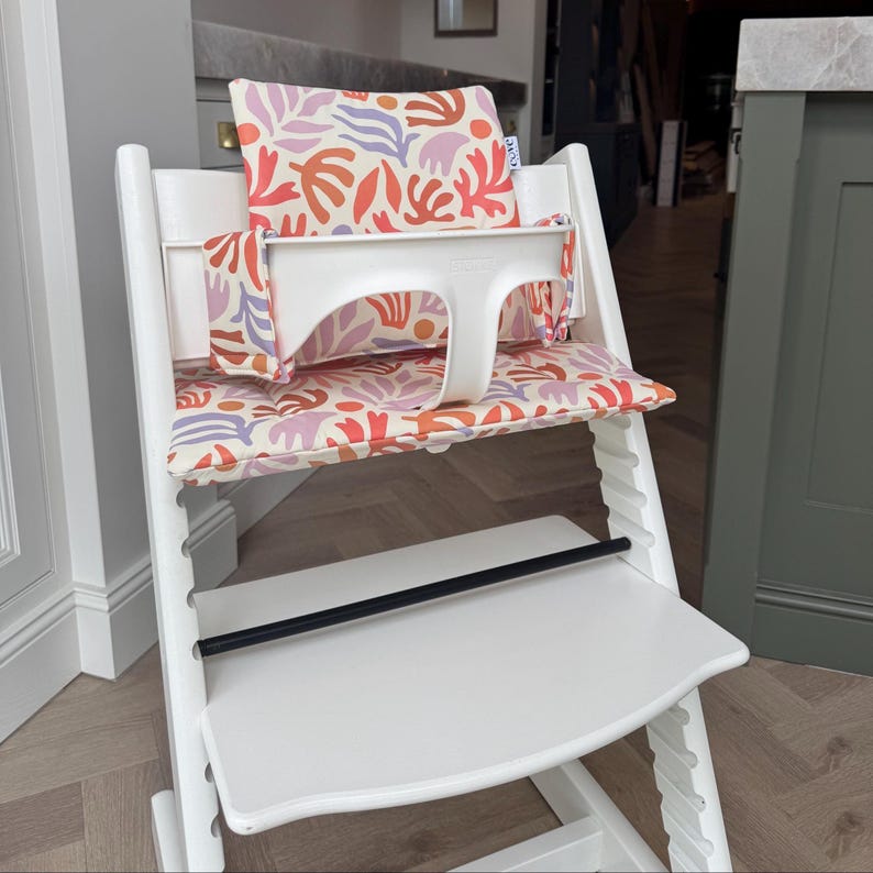 May include: A white wooden high chair featuring a removable white plastic safety bar and a cushioned seat pad and backrest. The fabric displays a repeating abstract pattern of coral, lavender, and orange organic shapes on a cream background.