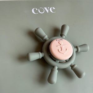 Puede incluir: Un mordedor gris para bebés con forma de timón de barco, con un centro rosa con un diseño de ancla. La palabra "cove" está impresa en el mordedor y el fondo.