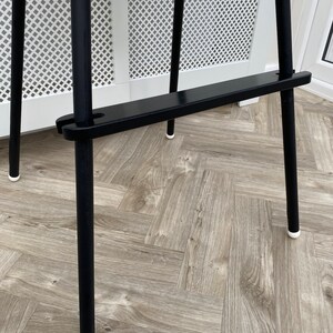 A black wooden footrest that is fitted on a toddlers  high chair legs. Accompanied with black leg wraps on the high chair legs. Placed on a wooden floor.